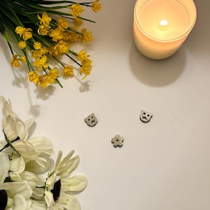 May include: A lit candle in a white glass jar, yellow flowers, and white flowers are arranged on a white surface. Three small wooden bear and flower-shaped embellishments are scattered on the surface.