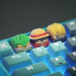 以下が含まれることがあります： アニメのキャラクターデザインが施されたカスタムキーキャップを備えたメカニカルキーボードのクローズアップ。キーキャップは、緑色の髪、麦わら帽子、金髪のキャラクターを描いています。キーボードは、白い文字が入ったティールキーと黒い背景です。