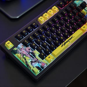 Puede incluir: Un teclado mecánico negro con teclas amarillas y negras. El teclado presenta el texto "Cyberpunk 2077" y teclas de acento coloridas. Las teclas tienen varios diseños y colores, incluyendo amarillo, verde y morado.