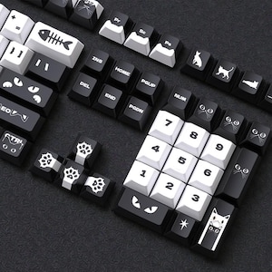 Puede incluir: Un conjunto de teclas de teclado en blanco y negro con diseños temáticos de gatos. Las teclas presentan caras de gato, huellas de patas y gráficos de espina de pescado. Las teclas están dispuestas sobre una superficie oscura.