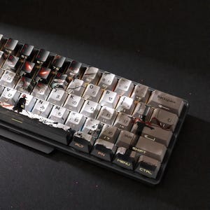 Puede incluir: Un teclado mecánico con teclas personalizadas que presenta un diseño en blanco y negro con caracteres japoneses y una figura de samurái. El teclado tiene un marco gris oscuro.