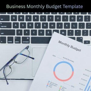 May include: A laptop keyboard with a pair of spectacles and a printed "Monthly Budget" document. The document features a pie chart with red and blue sections, along with a bar graph. The text "Business Monthly Budget Template" is at the top.