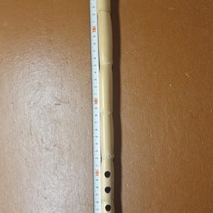May include: A light-colored bamboo flute with a copper-colored end cap. The flute has several finger holes and is approximately 12 inches long. A ruler is placed next to the flute for measurement.