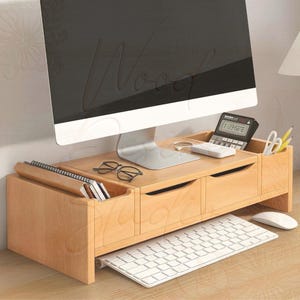 Puede incluir: Un soporte de monitor de madera de color claro con dos cajones, una calculadora y un portalápices. Un teclado y un ratón blancos están delante del soporte. Un monitor de ordenador se encuentra encima del soporte.