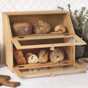 May include: A light-coloured wooden bread bin with two glass-fronted compartments. The top shelf holds various loaves of bread, while the bottom shelf displays sliced and whole loaves. The bin is open, revealing the contents.