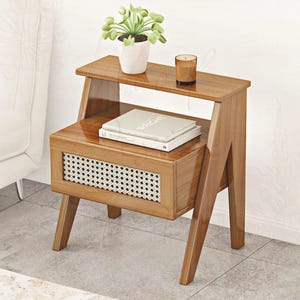 Puede incluir: Una mesa auxiliar de madera con un dise&ntilde;o de dos niveles. La repisa superior contiene una peque&ntilde;a planta en maceta y una vela. La secci&oacute;n inferior presenta un caj&oacute;n con un panel tejido y una pila de libros. La mesa es de madera marr&oacute;n claro.
