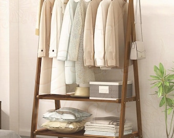 Puede incluir: Un perchero de madera en forma de A con una barra superior que sostiene varios abrigos y camisas de tonos claros. La unidad incluye dos estantes inferiores con una caja de almacenamiento, dos cojines decorativos y textiles doblados.