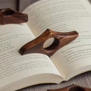 May include: Wooden thumb book page holders with a natural wood grain finish. The ergonomic design features a finger hole for comfortable reading. The book accessories are shown on the pages of an open book.