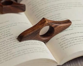 Puede incluir: Soportes de página de libro de pulgar de madera con un acabado natural de vetas de madera. El diseño ergonómico presenta un orificio para el dedo para una lectura cómoda. Los accesorios del libro se muestran en las páginas de un libro abierto.