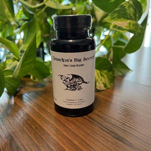 May include: A black bottle with a black cap, labeled "Grandpa's Big Secret Nitric Oxide Booster." The label features a black and white illustration of a winged creature. The bottle is on a wooden surface, with green plants in the background.