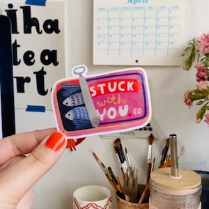 Pode incluir: Um autocolante com um design de lata de sardinhas com dois peixes azuis e o texto "Stuck with You .co" em rosa e branco. O autocolante é segurado em frente a uma parede com outros autocolantes e um calendário.