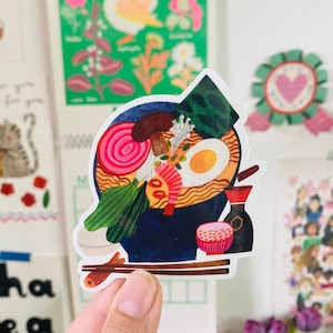 May include: A colourful sticker depicting a bowl of ramen with toppings such as egg, seaweed, and vegetables. The sticker also includes a small dish of rice, soy sauce, and chopsticks. The design is vibrant and detailed.