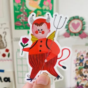 Könnte beinhalten: Ein Aufkleber mit einer Cartoon-Teufelsfigur in roter Kleidung, die einen Dreizack und eine Rose hält. Der Teufel hat Hörner, Flügel und einen Schwanz. Der Aufkleber wird von einer Hand gehalten, mit anderen dekorativen Bildern im Hintergrund.