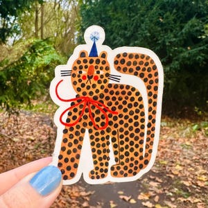 Könnte beinhalten: Ein Aufkleber mit einem Cartoon-Leoparden mit orangefarbenen Flecken und einer blauen Partyhütchen. Der Leopard hat eine rote Schleife und schwarze Schnurrhaare. Der Aufkleber wird vor einem verschwommenen Hintergrund aus Bäumen und Herbstlaub gehalten.