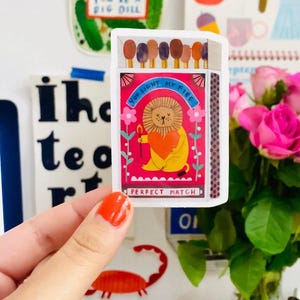 May include: A matchbox sticker with the text "YOU LIGHT MY FIRE" and "PERFECT MATCH", featuring a lion holding a heart. Other stickers include a jar of pickles and an apple. The image is bright and colourful.