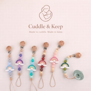 May include: A selection of baby pacifier clips with rainbow and cloud designs. The clips feature wooden beads, colourful silicone beads, and a wooden clip. The image includes the text "Cuddle & Keep" and the tagline "Made to cuddle. Made to keep."