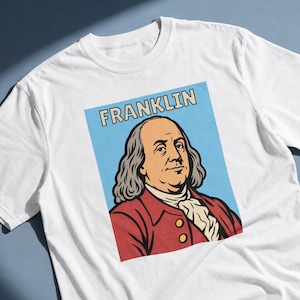 May include: A white short-sleeved t-shirt featuring a graphic print of Benjamin Franklin. The design shows a portrait of Franklin in a red coat against a light blue background with the word FRANKLIN printed in bold letters above the portrait.