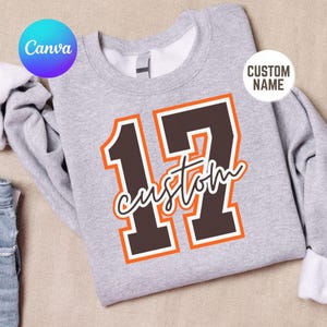 May include: A light gray crewneck sweatshirt featuring a large dark brown number 17 with a bright orange outline. The word custom is written in a white cursive script font across the center of the number 17. The garment has long sleeves.