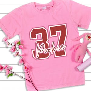 May include: A light pink short-sleeved t-shirt featuring a large dark red number 37 with a white outline. Below the number, the word Pookie is printed in a white cursive script font. The shirt is displayed with a pink plush toy and pink flowers.