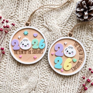 May include: Two round wooden ornaments with white borders and twine loops. Each features a smiling dumpling character, the year 2026 in pastel numbers, and the text THE YEAR OF THE SQUISHIES. Small pastel stars and hearts decorate the light wood surface.