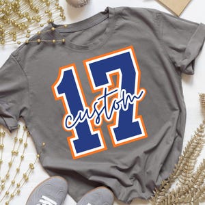 May include: A charcoal gray short-sleeve t-shirt featuring a large navy blue number 17 with a white and orange outline. The word custom is written in a white cursive script font across the center of the number 17 on the front of the shirt.