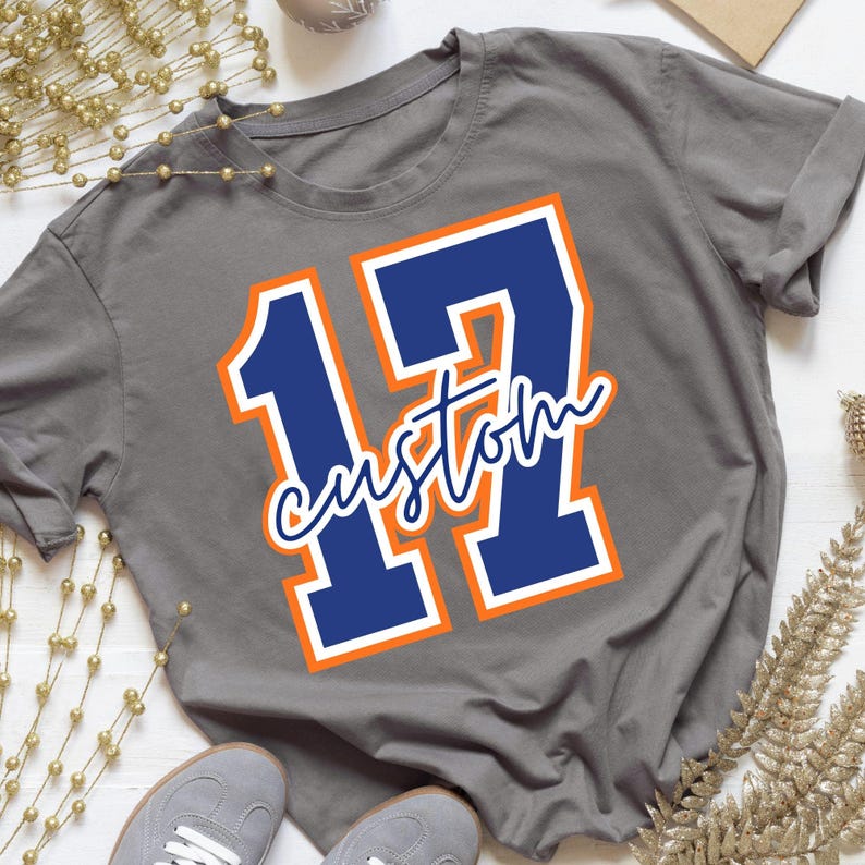 May include: A charcoal gray short-sleeve t-shirt featuring a large navy blue number 17 with a white and orange outline. The word custom is written in a white cursive script font across the center of the number 17 on the front of the shirt.