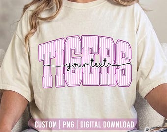 May include: A cream-colored short-sleeve t-shirt featuring the word TIGERS in large, purple-outlined block letters with a vertical pink striped pattern. The words your text are written in a black cursive script across the center of the design.