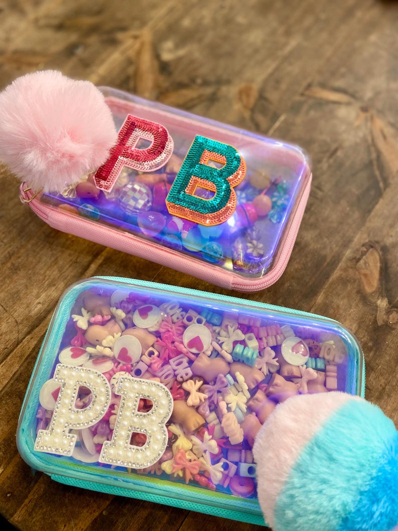 May include: Two clear pencil cases with colorful contents and the initials "PB". One case is pink with a pink pom-pom, the other is teal with a blue and pink pom-pom. The cases are filled with small decorative items.