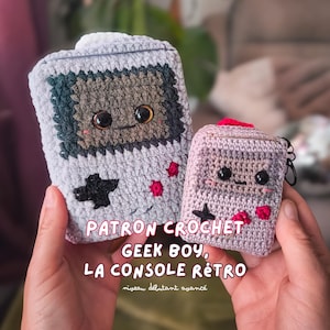 May include: Two crocheted cases resembling retro handheld game consoles. The larger case is gray with a brown screen and black cross-shaped buttons. The smaller case is pink with a red top and black accents. The text "PATRON CROCHET, GEEK BOY, LA CONSOLE RÉTRO" is visible.