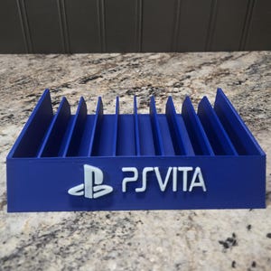 May include: A blue plastic storage rack for game cartridges featuring ten vertical slots and the white PlayStation logo followed by the text PS VITA embossed on the front face of the base. The unit has a rectangular shape with a flat bottom.