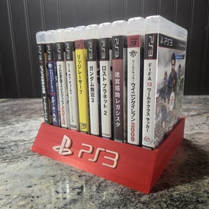 May include: A red plastic display stand featuring the white PS3 logo, holding a row of ten PlayStation 3 video game cases. The game spines display various Japanese titles including FIFA 13, Winning Eleven 2009, Lost Planet 2, Gundam Musou 3, and Ridge Racer 7.