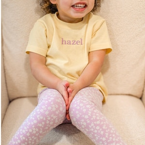 May include: A light yellow t-shirt with the name "hazel" embroidered in lavender. The child is wearing light purple leggings with a white floral pattern. The outfit is for a child.