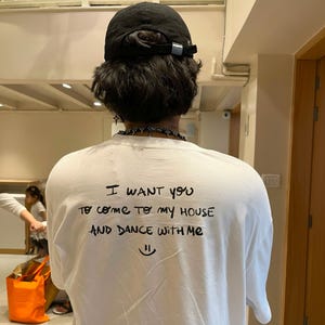 Puede incluir: Camiseta blanca con el texto escrito a mano "I want you to come to my house and dance with me" y una carita sonriente. La persona lleva una gorra negra y un collar.