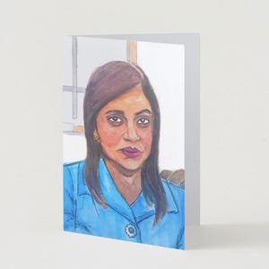 May include: A watercolor portrait on a greeting card. The subject is a woman with dark hair and a blue collared shirt. The card is white and folded, with the portrait on the front.