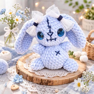 May include: A light blue crocheted bunny doll with large blue eyes and white bows. The handmade plush toy has a stitched face and an 'X' on its chest. It sits on a wooden slice with a lace doily, surrounded by flowers and yarn.