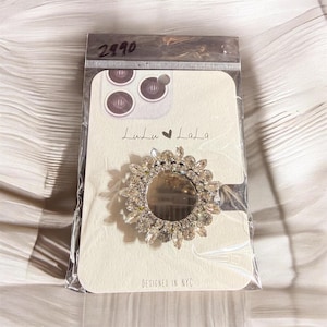 May include: A clear plastic package containing a phone grip. The phone grip is a silver-colored, circular design with a mirrored center and a surrounding array of clear, sparkling crystals. The card behind the grip has the text "Lulu LaLa" and "DESIGNED IN NYC".