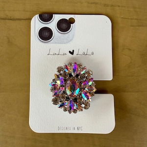May include: A bedazzled phone grip with an iridescent, oval-shaped center stone surrounded by smaller, colorful gems. The grip is attached to a white card with the text "Lulu Lala" and "Designed in NYC".