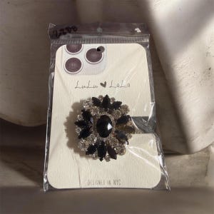 May include: A black and silver rhinestone brooch in a floral design, set on a white card with the text "Lulu LaLa" and "Designed in NYC". The brooch has a large black center stone.