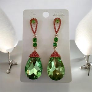 May include: A pair of long dangle earrings featuring a teardrop-shaped green crystal at the base, topped with a red crystal cap. Above this, two small green crystals are set in a red frame, leading to a red crystal loop at the top of each earring.