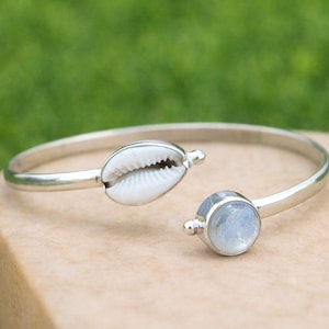 May include: A silver-toned cuff bracelet featuring a white seashell and a round, light blue gemstone. The open design allows for easy wear. The bracelet is displayed on a light brown surface against a blurred green background.