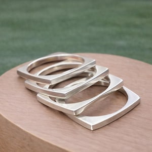 May include: Four silver square bangles stacked on a wooden surface. The bangles have a minimalist design with rounded inner edges and sharp outer corners. The background is a blurred green.