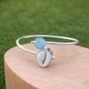 May include: A silver-toned bracelet featuring a light blue gemstone and a white seashell. The bracelet has an open design with the gemstone and seashell as focal points. The bracelet is displayed on a light brown surface against a green background.