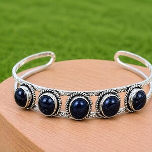 May include: A silver cuff bracelet featuring five oval-shaped, dark blue, glittery cabochon stones. The stones are set in twisted rope-style metal frames and connected by two parallel silver bands with a textured, dotted surface finish.