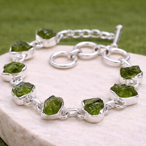 May include: A silver bracelet featuring a series of vibrant green peridot stones. Each stone is set in a silver bezel, connected by silver links. The bracelet has a toggle clasp. The background is a light-colored surface with a green backdrop.