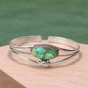 Puede incluir: Brazalete de plata con una piedra turquesa. La piedra turquesa ovalada está engastada en un bisel de plata con un diseño de cuerda trenzada. El brazalete tiene un elemento decorativo de plata en la parte inferior.