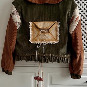 Pode incluir: Casaco de veludo cotelê verde-oliva e castanho com um design único. Apresenta gola e mangas castanhas, e um bolso decorativo em forma de envelope com acabamento em renda. Um charme de cogumelo pende da bainha.