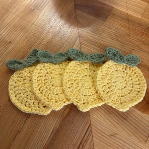 May include: Four handmade crochet coasters in a lemon slice design. The coasters are yellow with a green leaf detail at the top. The coasters are arranged on a wooden surface.