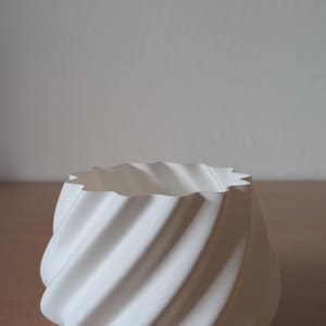 May include: A white, spiral-shaped ceramic planter with a textured surface. The planter has a wavy top edge and sits on a wooden surface. The design is modern and minimalist, suitable for small plants or decorative use.