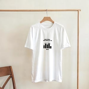 Puede incluir: Una camiseta blanca con un diseño gráfico negro. El diseño incluye el texto "NICE STORY, NOW SHOW ME THE DATA" encima de una ilustración de un gráfico de barras. La camiseta está colgada en una percha de madera.