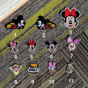 Puede incluir: Una colección de once parches bordados con varios personajes de Disney. Los parches incluyen Mickey Mouse, Minnie Mouse, Tigger, el tarro de miel de Winnie the Pooh, Piglet, Donald Duck y Pluto. Cada parche tiene un diseño detallado y acentos coloridos.